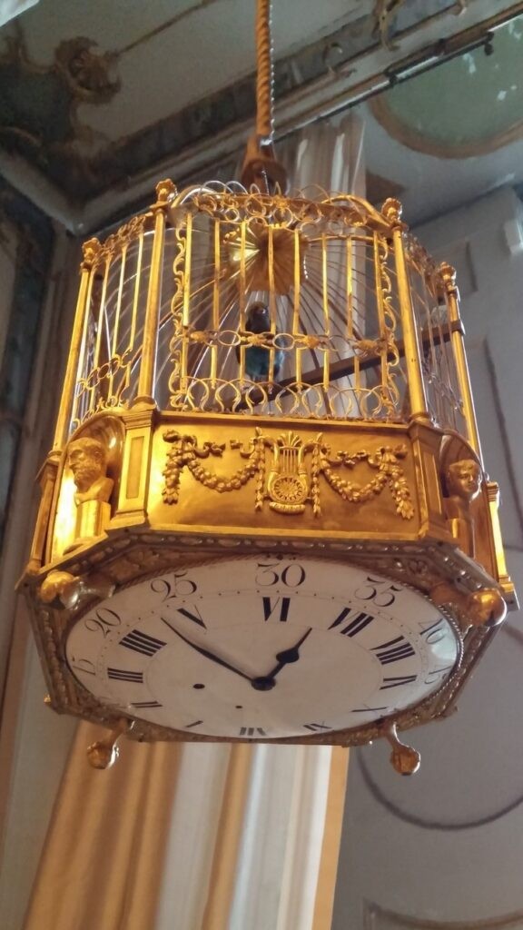 An elaborate, gold-plated birdcage hanging from the ceiling, with a clock face you can see from the bottom. A custom clock, one of a pair that was a gift from Marie Antoinette to her sister Mary Caroline